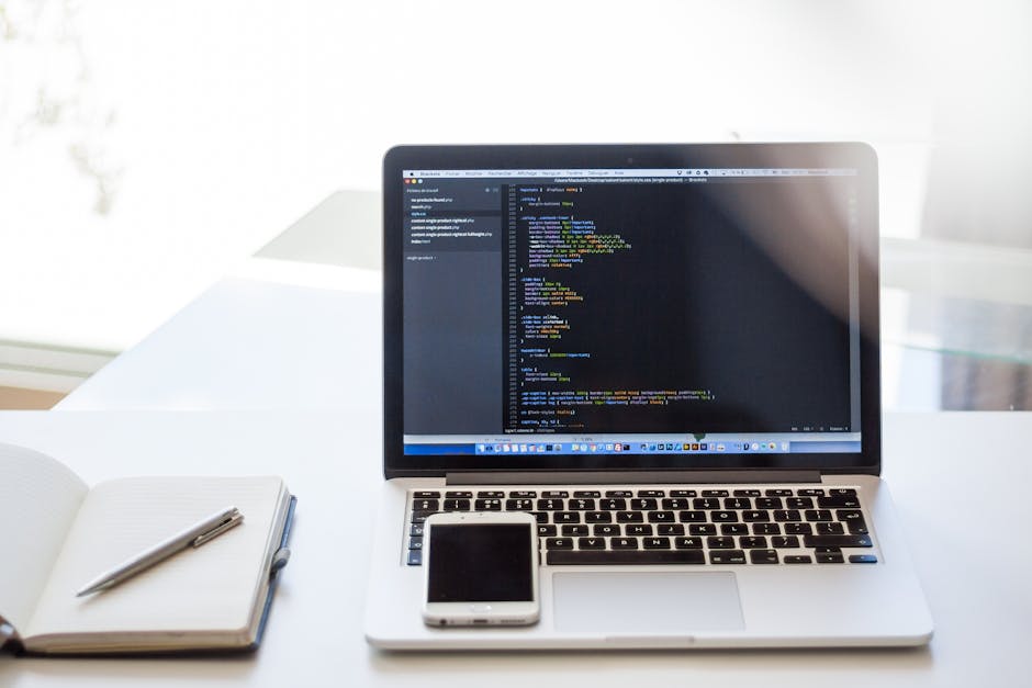 A close-up view of a laptop on a white desk showing lines of code on the screen, with an open notebook and a pen placed beside it, and a smartphone resting on the laptop's trackpad. The background fea