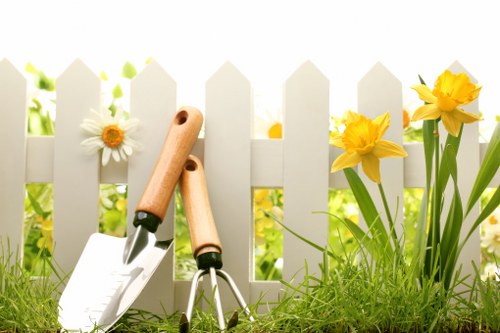 A close-up view of a small section of a garden in Colindale, showing a white picket fence with evenly spaced vertical slats in the background, partly shaded by sunlight. In the foreground, there is a lush, green grass lawn with fine, dense blades, alongside a few blooming yellow daffodils with tall, green leafy stems. To the left of the flowers, gardening tools including a trowel and a hand rake with light wooden handles are partially embedded in the soil, suggesting ongoing gardening work. The environment appears well-maintained and vibrant, with natural daylight casting soft shadows, indicating a clear, mild weather day ideal for outdoor gardening activities. This scene exemplifies a healthy, landscaped front or backyard typical of residential outdoor spaces in Colindale, supporting professional gardening and lawn care services provided by Gardening Services Colindale.